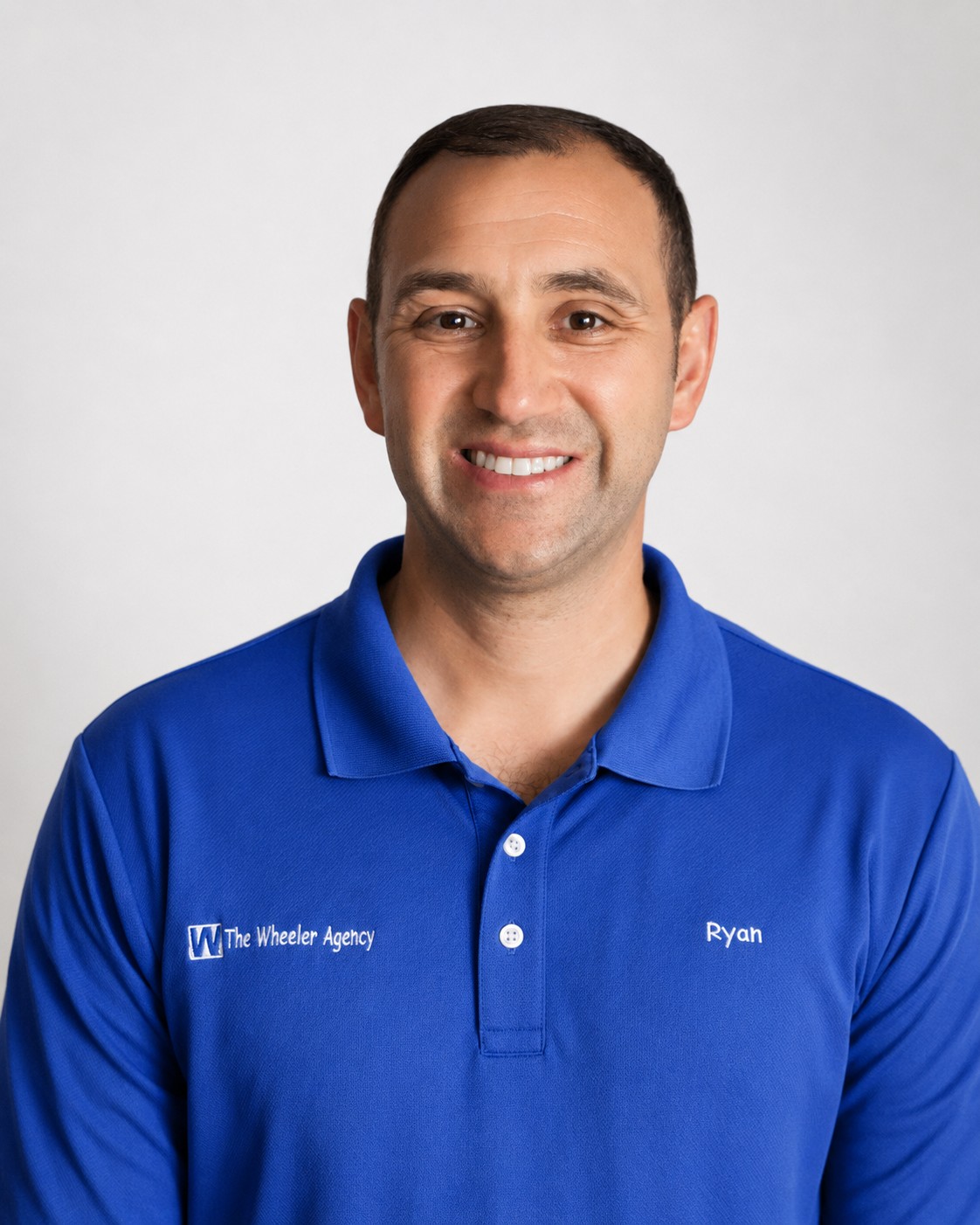 A man smiles while wearing a blue polo shirt with a logo and his name embroidered on it.